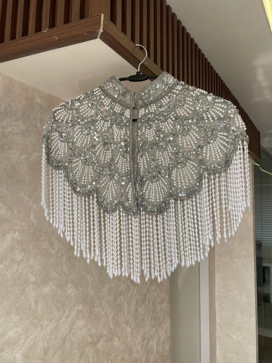 A silver beaded cape hangs on a hanger against a beige wall. The cape has a high collar and is decorated with intricate beadwork and tassels.