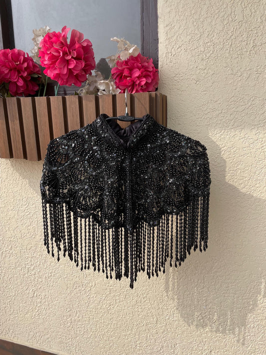A silver beaded cape hangs on a hanger against a beige wall. The cape has a high collar and is decorated with intricate beadwork and tassels.
