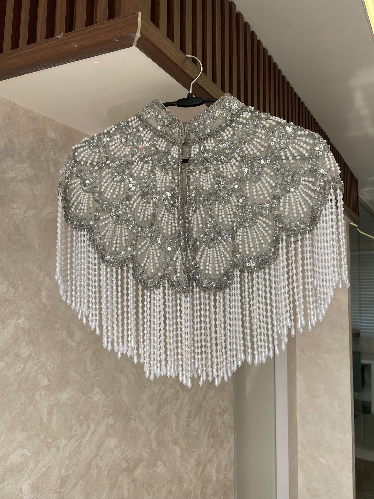 A silver beaded cape hangs on a hanger against a beige wall. The cape has a high collar and is decorated with intricate beadwork and tassels.