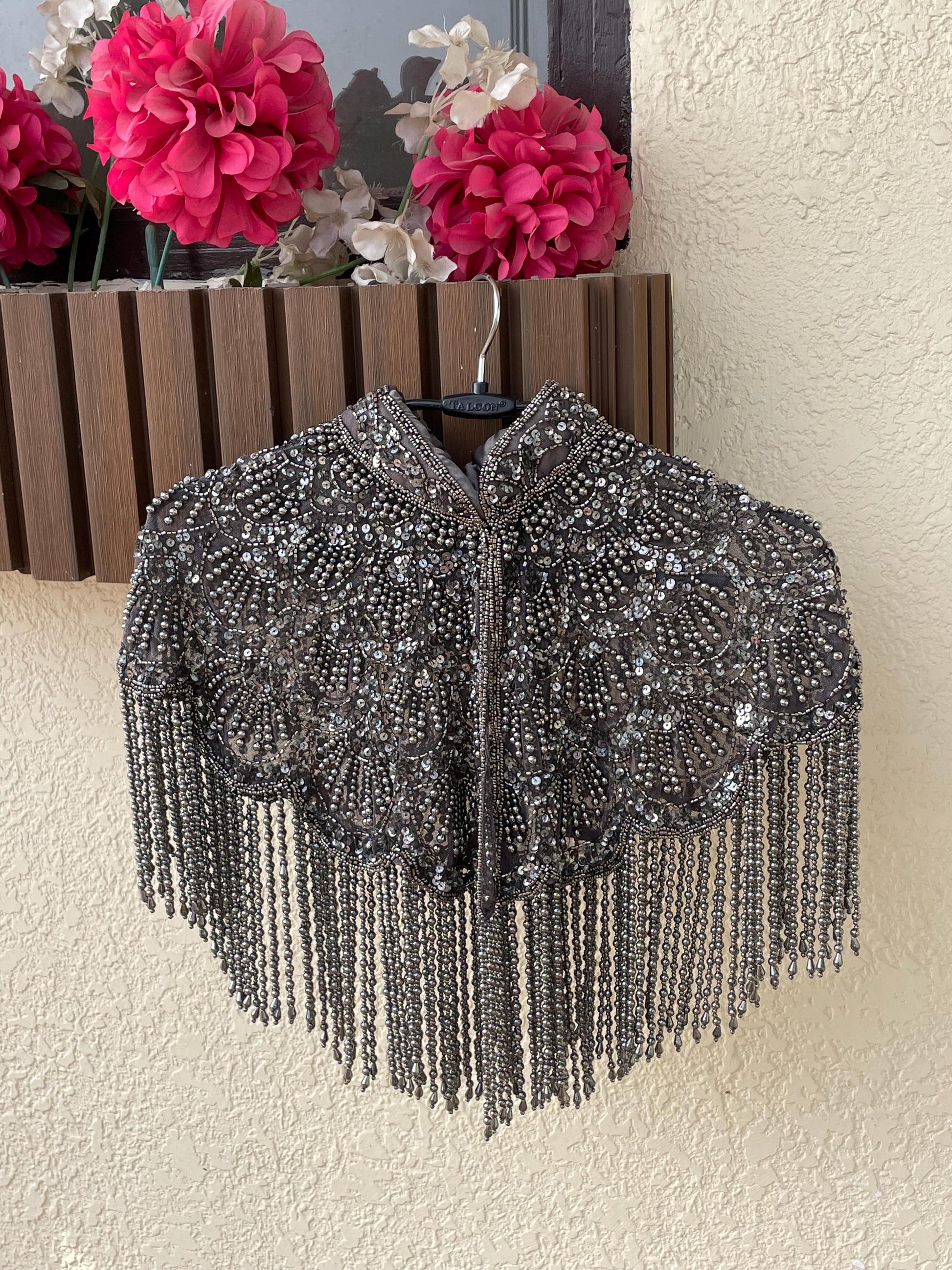 A silver beaded cape hangs on a hanger against a beige wall. The cape has a high collar and is decorated with intricate beadwork and tassels.