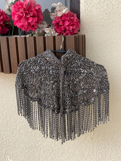 A silver beaded cape hangs on a hanger against a beige wall. The cape has a high collar and is decorated with intricate beadwork and tassels.