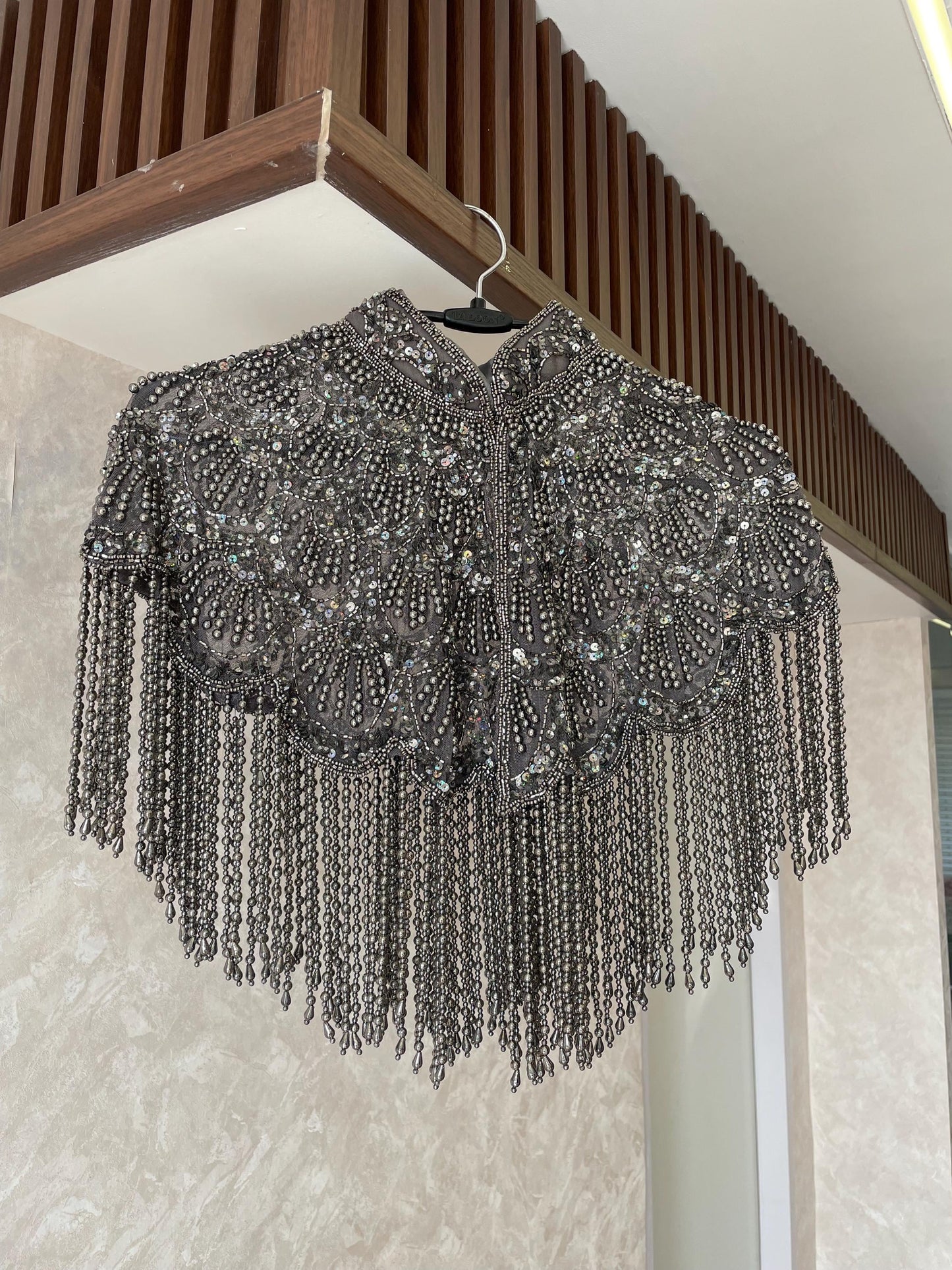 A silver beaded cape hangs on a hanger against a beige wall. The cape has a high collar and is decorated with intricate beadwork and tassels.
