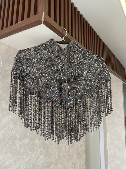 A silver beaded cape hangs on a hanger against a beige wall. The cape has a high collar and is decorated with intricate beadwork and tassels.